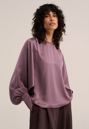 Femme aux cheveux bouclés foncés vêtue d'un chemisier mauve ample à manches longues avec poignets froncés et d'un pantalon sombre sur un fond uni.