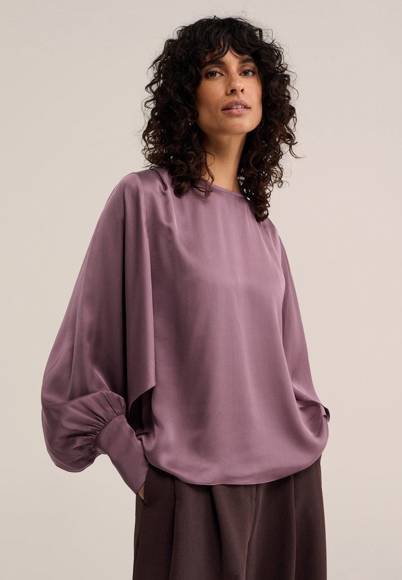Femme aux cheveux bouclés foncés vêtue d'un chemisier mauve ample à manches longues avec poignets froncés et d'un pantalon sombre sur un fond uni.