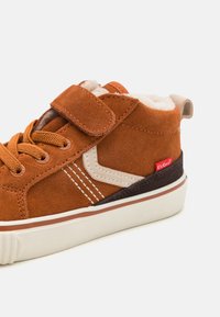 Zapatilla de ante marrón con cordones y tira de velcro, con forro crema, acentos beige en contraste y una etiqueta de logo roja. Suela de goma.