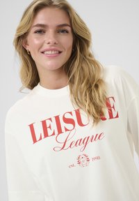 White oversized t-shirt with red “LEISURE League” text. Features a round neckline and soft, smooth fabric. Casual design with minimal details.