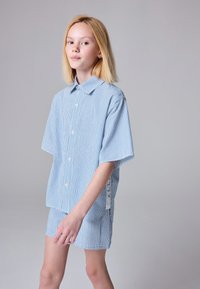 Plava i bijela prugasta košulja s kratkim rukavima i kopčanjem na gumbe, u kombinaciji s pripadajućim kratkim hlačama. Materijal izgleda lagano i ležerno.