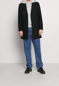 Černý kabát z vlněné směsi se třemi knoflíky, nošený přes šedou mikinu s kapucí, v kombinaci s modrými džínami a černými šněrovacími botami.