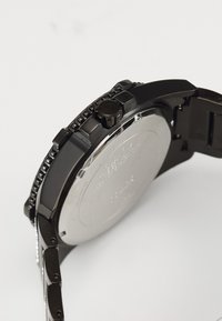Svart klocka med ett stort metallfall, texturerad bezel och en framträdande krona. Baksidan är slät silverfärgad med ingraverade detaljer.