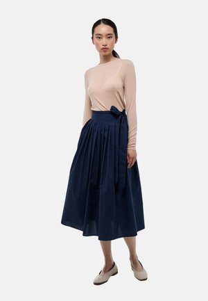 Femme portant un haut à manches longues beige rentré dans une jupe midi plissée bleu marine avec un nœud sur le côté, associée à des chaussures plates beiges.