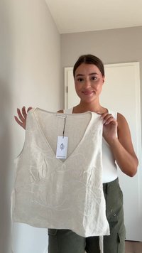 Top senza maniche color crema con ricami floreali, scollatura a V e lacci ai lati. Il tessuto sembra leggero e testurizzato.