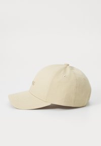 Beige baseball cap med en buet skjerm, stofftekstur og broderte detaljer på framsiden. Har ventilasjonslufter på toppen.