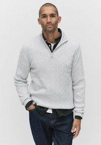 Ljusgrå kabelstickad tröja med hög krage och fjärdedeldragkedja, lager-på-lager över en mörkgrön skjorta, kombinerad med mörkblå jeans.