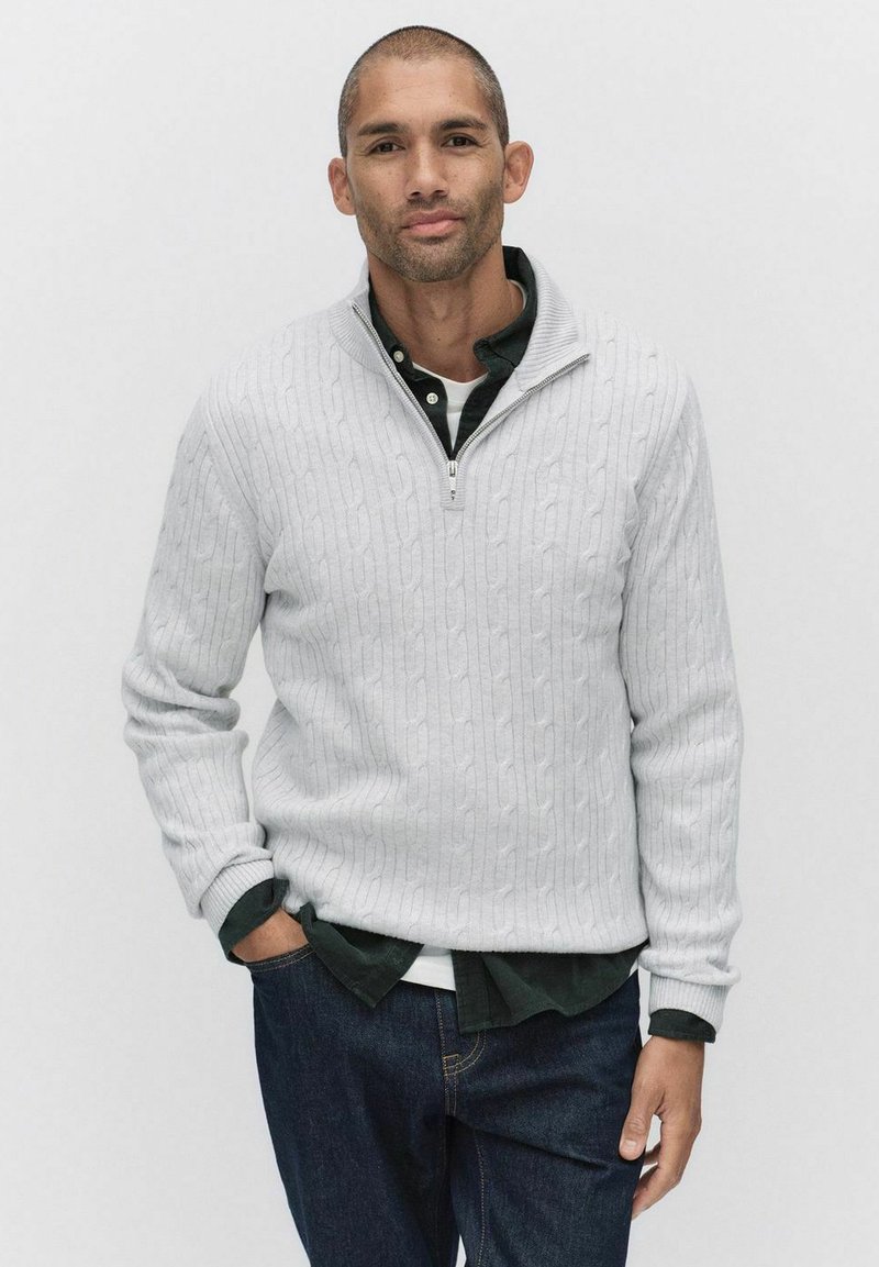 Ljusgrå kabelstickad tröja med hög krage och fjärdedeldragkedja, lager-på-lager över en mörkgrön skjorta, kombinerad med mörkblå jeans.