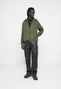 Μπουφάν ελιάς με φερμουάρ και υφασμένα μανίκια, συνδυασμένο με μαύρο δερμάτινο παντελόνι cargo. Το σύνολο ολοκληρώνεται με μαύρα αθλητικά παπούτσια.