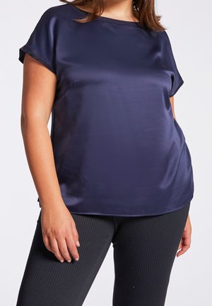 Frau trägt eine marineblaue Satinhose mit kurzen Ärmeln und strukturierte schwarze Hose, steht mit einer Hand entspannt an der Seite.