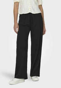 Pantalons larges noirs en tissu léger, dotés d'une taille élastique avec un cordon de serrage et d'une coupe décontractée. Associés à des baskets blanches.