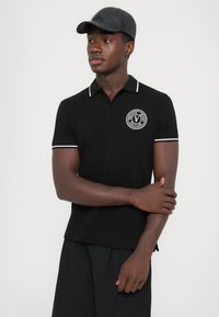 Bărbat îmbrăcat cu un tricou polo negru cu dantelă albă și logo-ul Versace Jeans Couture, purtând o șapcă neagră și pantaloni negri, stând în fața unui fundal simplu.