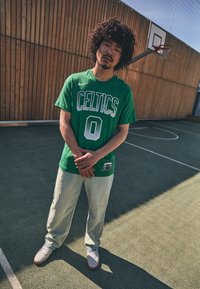 Green Celtics t-shirt with white lettering, light green pants, and grey sneakers with pink accents, standing on a basketball court.