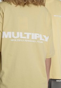 Gelbes Baumwoll-T-Shirt mit großem weißem Textaufdruck "MULTIPLY" und "MULTIPLY APPAREL CLUB" auf der Rückseite; lockerer Schnitt, weiche Textur.