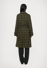 Pikk ruuduline vihmamantel rohelises ja mustas, millel on vöökohal rihm, laiad kraed ja pikad varrukad, sirge lõige ja tekstuuriga kangas.