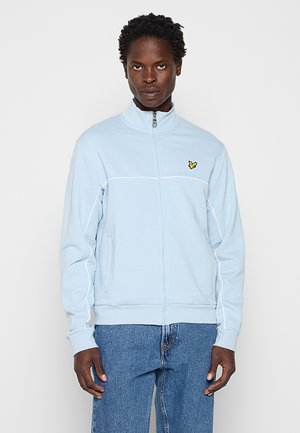 Junger Mann trägt eine hellblaue Jacke mit Reißverschluss, weißen Paspeln und einem kleinen gelben Vogel-Logo, kombiniert mit blauen Jeans.