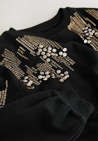 Svart sweatshirt med guld- och iridescerande paljetter arrangerade i ett dekorativt mönster. Ribbad muddar och halsringning förbättrar textur och passform.