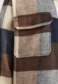 Gros plan sur une poche à rabat boutonnée sur une veste en laine à carreaux marron, beige et bleu.