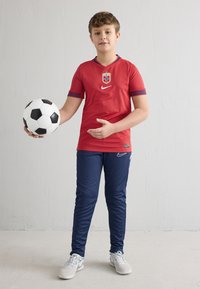 Raudoni „Nike“ futbolo marškinėliai su mėlynais akcentais ir emblema, derinami su tamsiai mėlynomis sportinėmis kelnėmis. Vaikas laiko juodai baltą futbolo kamuolį.