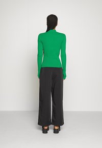 Pull à col roulé côtelé vert associé à un pantalon noir à jambes larges. La tenue présente une texture lisse et une coupe ajustée. Chaussures noires épaisses.