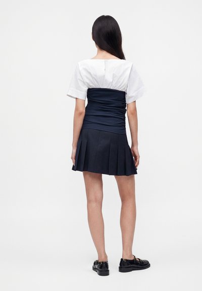 Femme aux longs cheveux foncés portant un haut blanc à manches courtes avec un corset bleu marine ajusté et une jupe plissée bleu marine, debout, tournée de dos.
