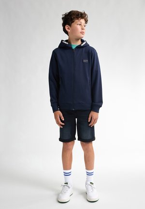 Sweat à capuche zippé bleu marine avec une capuche bordée de blanc, associé à un short en denim retroussé. Des baskets blanches avec des accents verts et bleus complètent la tenue.