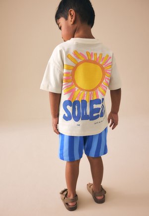 Barn iført en hvid t-shirt med en stor gul sol og blå tekst "Soleil" på ryggen, blå stribede shorts og brune sandaler.