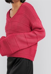 Rosa gestrickter Pullover mit V-Ausschnitt, der strukturierte Einsätze und weite Ärmel aufweist. Der gerippte Saum fügt Details hinzu und wird mit schwarzen Hosen kombiniert.