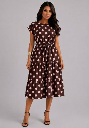 Femme portant une robe midi marron à pois roses, manches courtes, taille nouée et sandales noires à talons hauts sur un fond uni.