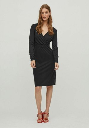 Black wrap dress with long sleeves, knee-length hem, and a smooth, stretchy texture; paired with red strappy sandals.