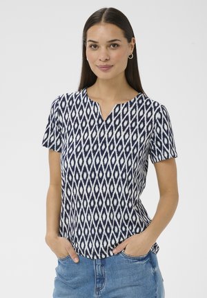 Woman with long dark hair wearing gold hoop earrings, a navy and white patterned short-sleeve top, and blue jeans with hands in pockets.