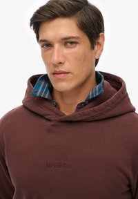 Felpa con cappuccio bordeaux con finitura testurizzata e logo ricamato. Sovrapposta a una camicia a quadri blu visibile all'colletto.