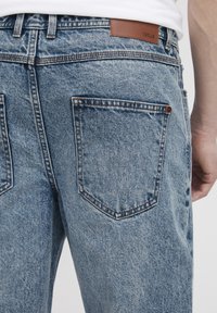 Lyseblå denimjeans med avslappet passform, med to baklommer, kontrastsøm og en brun skinnmerke ved midjen.