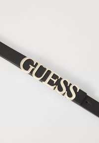 Čierny kožený opasok s výrazným zlatým kovovým logom "GUESS" ako stredobodom. Hladká textura s dierkami pre prispôsobenie veľkosti.