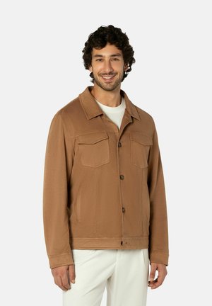 Homme portant une veste marron boutonnée avec des poches poitrine par-dessus une chemise blanche et un pantalon blanc, souriant devant un fond uni.