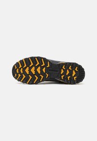 Semelle de chaussure New Balance noire et orange avec des crampons profonds et robustes pour une bonne adhérence sur diverses surfaces, présentée sur un fond clair uni.