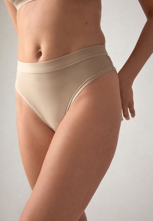 Vrouw met hoog opgesneden beige ondergoed, staand tegen een effen neutrale achtergrond, waarbij torso en bovenbenen zichtbaar zijn.