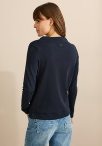 Mörkblå långärmad polo med krage, slät textur och en liten logotyp på baksidan, i kombination med ljusblå denimjeans.