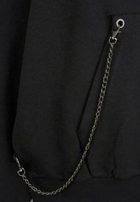 Czarna bluza z krótką kieszenią, ozdobiona metalowym łańcuszkiem przymocowanym za pomocą zapinki, wykonana z miękkiego materiału o gładkiej fakturze.