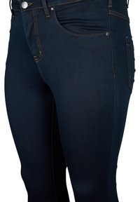 Jeans de mezclilla azul oscuro con costuras naranjas, un diseño estándar de cinco bolsillos y una silueta ajustada. Cuenta con un cierre frontal de botón y cremallera.