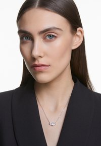 Colar de corrente prateada com dois destaques de gemas claras, usado sobre uma jaqueta preta, realçando o seu design simples e elegante.