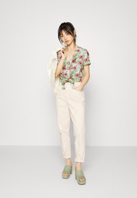 Blusa floral de mangas curtas com padrões verdes e rosas, atada na cintura; calças de cintura alta em cor creme; mules verde em plataforma.