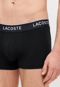 Czarne bokserki wykonane z elastycznej bawełny, z granatowym pasem z białym napisem "LACOSTE". Dopasowany krój, gładka tekstura.