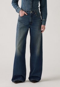 Jean en denim bleu foncé à jambe large et taille haute, avec des poches avant et un léger délavage sur les cuisses pour une texture.