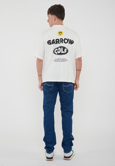 T-shirt blanche à manches courtes avec le texte graphique "BARROW GOLF" en noir, un logo rond jaune, associé à un jeans en denim bleu et des baskets blanches.