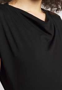 Gros plan d'une personne portant un haut noir avec un col lâche et drapé, avec des cheveux bouclés visibles sur le bord.