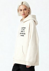 Felpa con cappuccio beige chiaro con tasca frontale, con testo nero che dice "A volte tutto ciò che ti serve sono 100 milioni di dollari." Tessuto morbido.