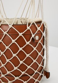 Sac seau en cuir marron avec haut à cordon en tissu beige, recouvert d’un filet en corde nouée de couleur crème et d’une bandoulière tressée.