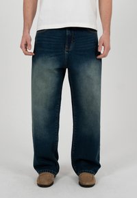 Bărbat purtând blugi albaștri deschiși, largi, mocasini bej și o cămașă albă, stând pe un fundal gri deschis.