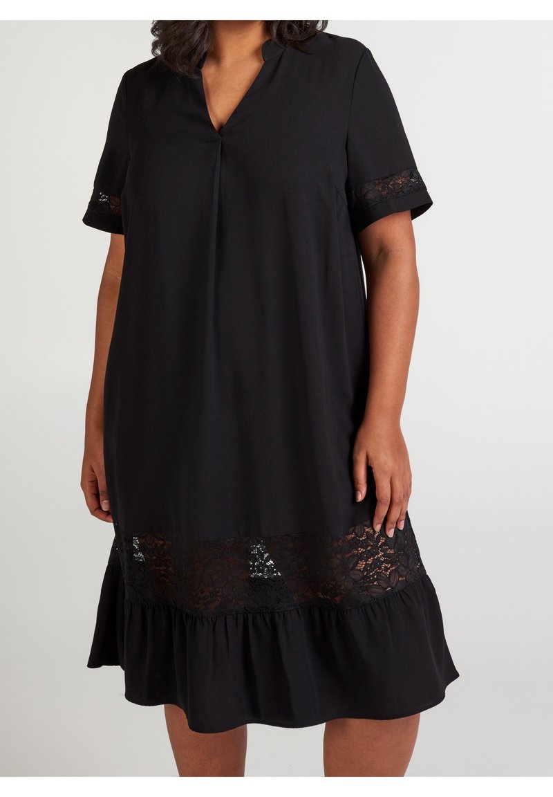 Femme portant une robe noire à manches courtes avec une finition en dentelle aux manches et à l'ourlet, debout devant un fond clair uni.